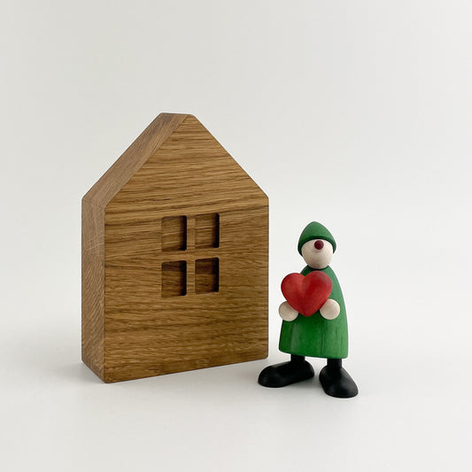 Ein Holz Dekohaus in Form eines Hauses mit vier quadratischen Fenstern steht auf einem schlichten weißen Hintergrund. Neben dem Haus befindet sich eine kleine Figur mit einem grünen, kegelförmigen Körper, einem weißen, runden Kopf und einem grünen, spitzen Hut. Die Figur hält eine rote Herzform vor sich.