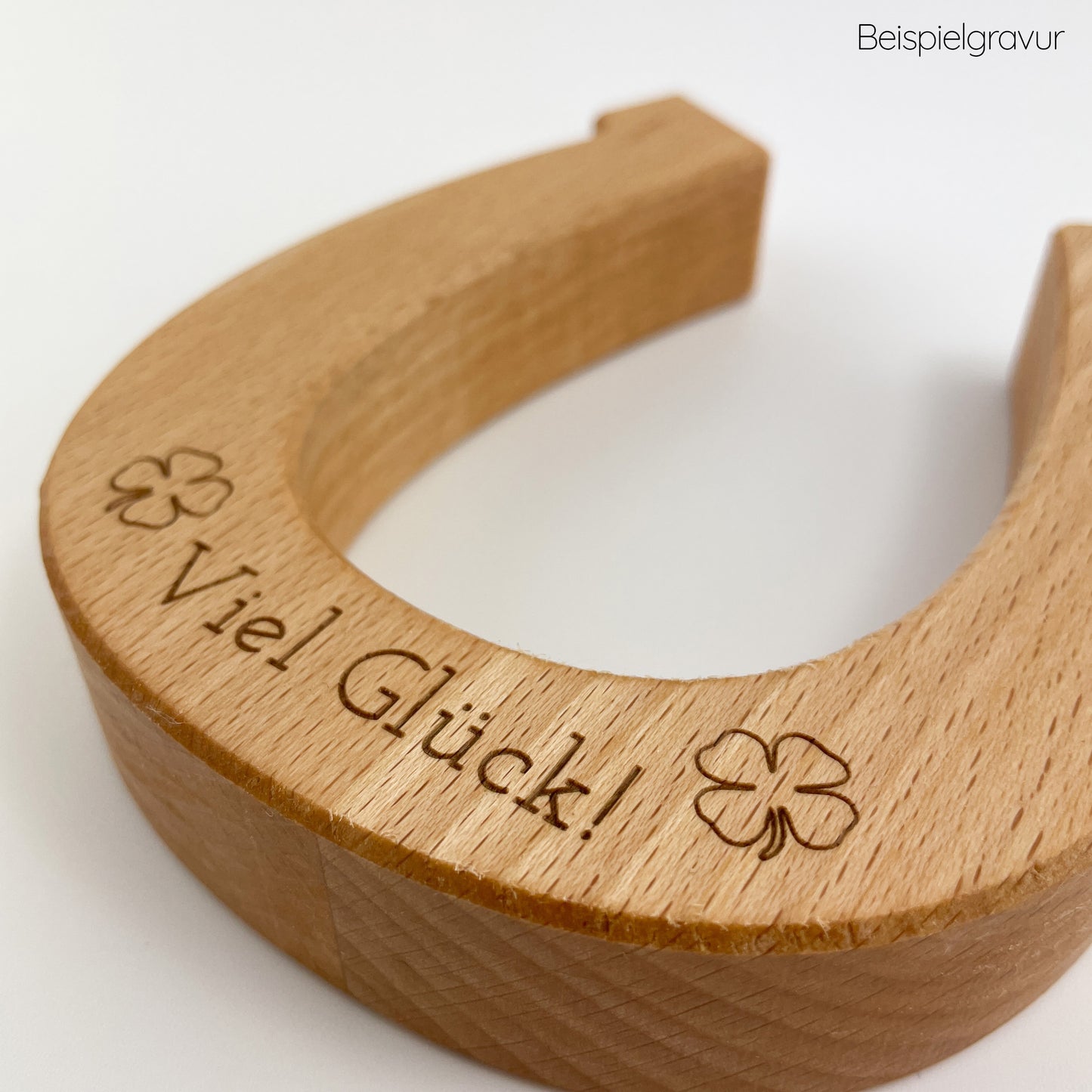 Detailansicht vonHufeisen aus Holz - Hugo, mit dem eingravierten Text “Viel Glück!” und Kleeblatt-Symbol auf einem weißen Untergrund liegend. Das Hufeisen zeigt die natürliche Maserung des Holzes und die warme Farbe sowie die präzise Gravur verleihen dem Objekt einen charmanten Charakter.