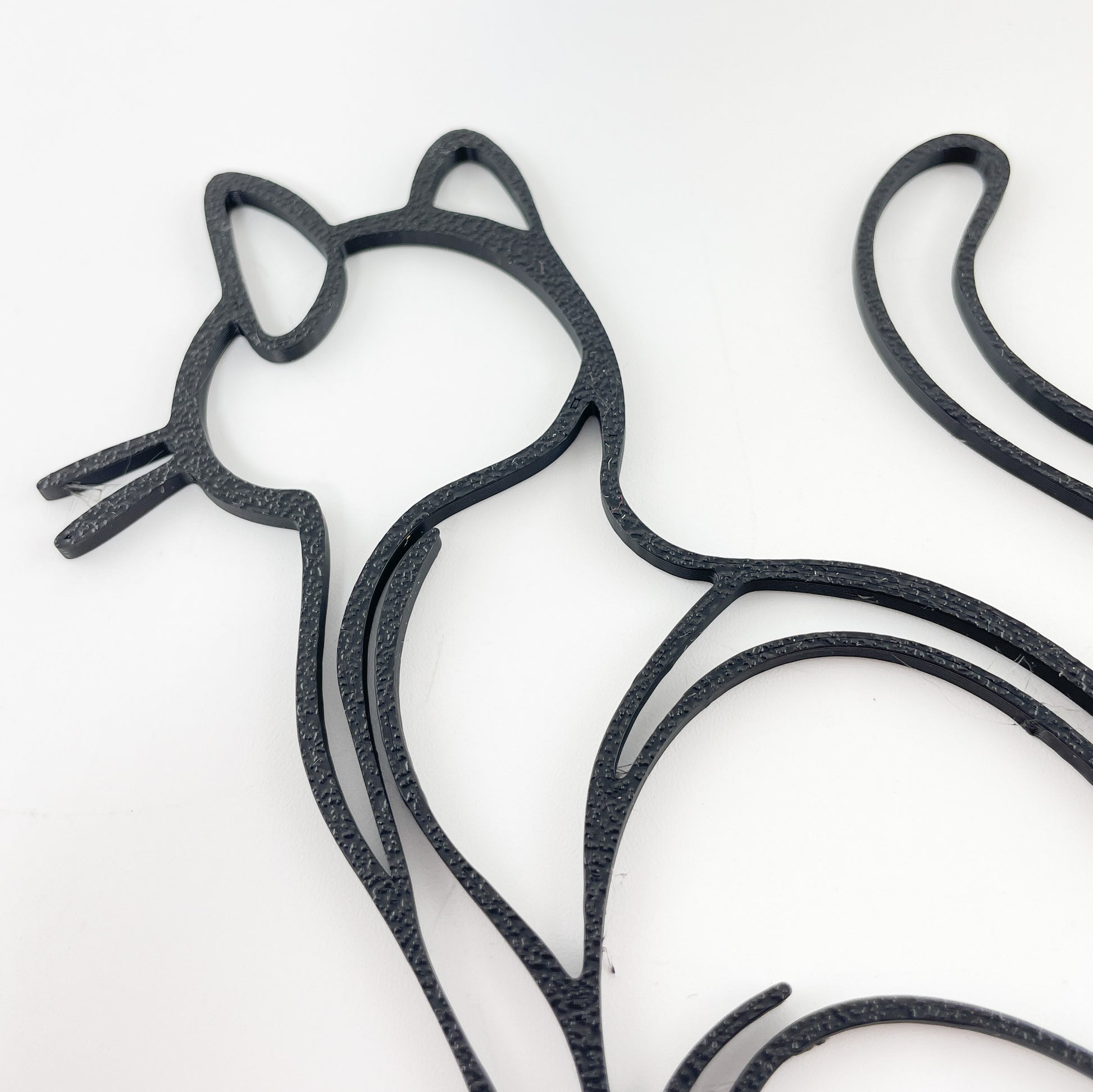 Detailaufnahme einer Katze von hinten, sitzend, mit aufgerichtetem Schwanz im Stile einer Linienzeichnung aus schwarzem Kunststoff vor weißem Hintergrund. Zu sehen ist die leichte Oberflächenstruktur und die Katze vom Kopf bis zur Hüfte.