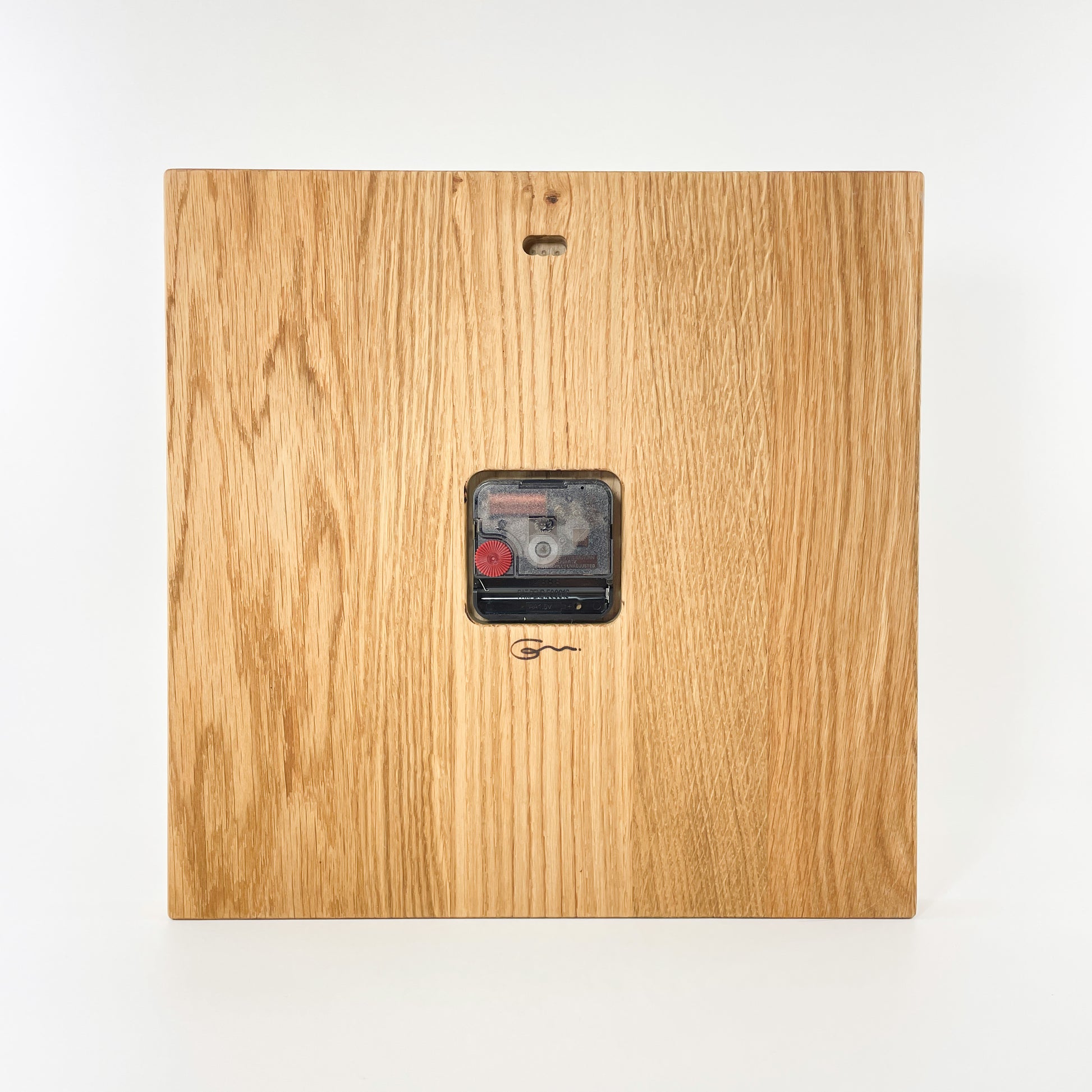Rückansicht der eckigen Wanduhr aus Holz mit römischen Ziffern auf der Vorderseite. Man sieht mittig platziert das eingelassene Uhrwerk und darunter das dezente Logo der Goertz Möbelmanufaktur Wismar. Oben ist ein Loch für die Aufhängung vorgesehen.