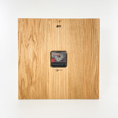 Rückansicht der eckigen Wanduhr aus Holz mit römischen Ziffern auf der Vorderseite. Man sieht mittig platziert das eingelassene Uhrwerk und darunter das dezente Logo der Goertz Möbelmanufaktur Wismar. Oben ist ein Loch für die Aufhängung vorgesehen.