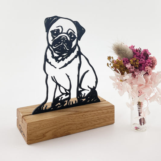 Linienzeichnung von einem Mops, die 3D-gedruckt als Steckbild in einem Halter aus massivem Eichenholz leicht schräg vor weißem Hintergrund steht. Rechts daneben steht ein kleiner Trockenblumenstrauß in Rosatönen in einer Glasvase. Links am Stirnende des Kartenhalters ist das eingebrannte Logo der Goertz Möbelmanufaktur zu sehen.