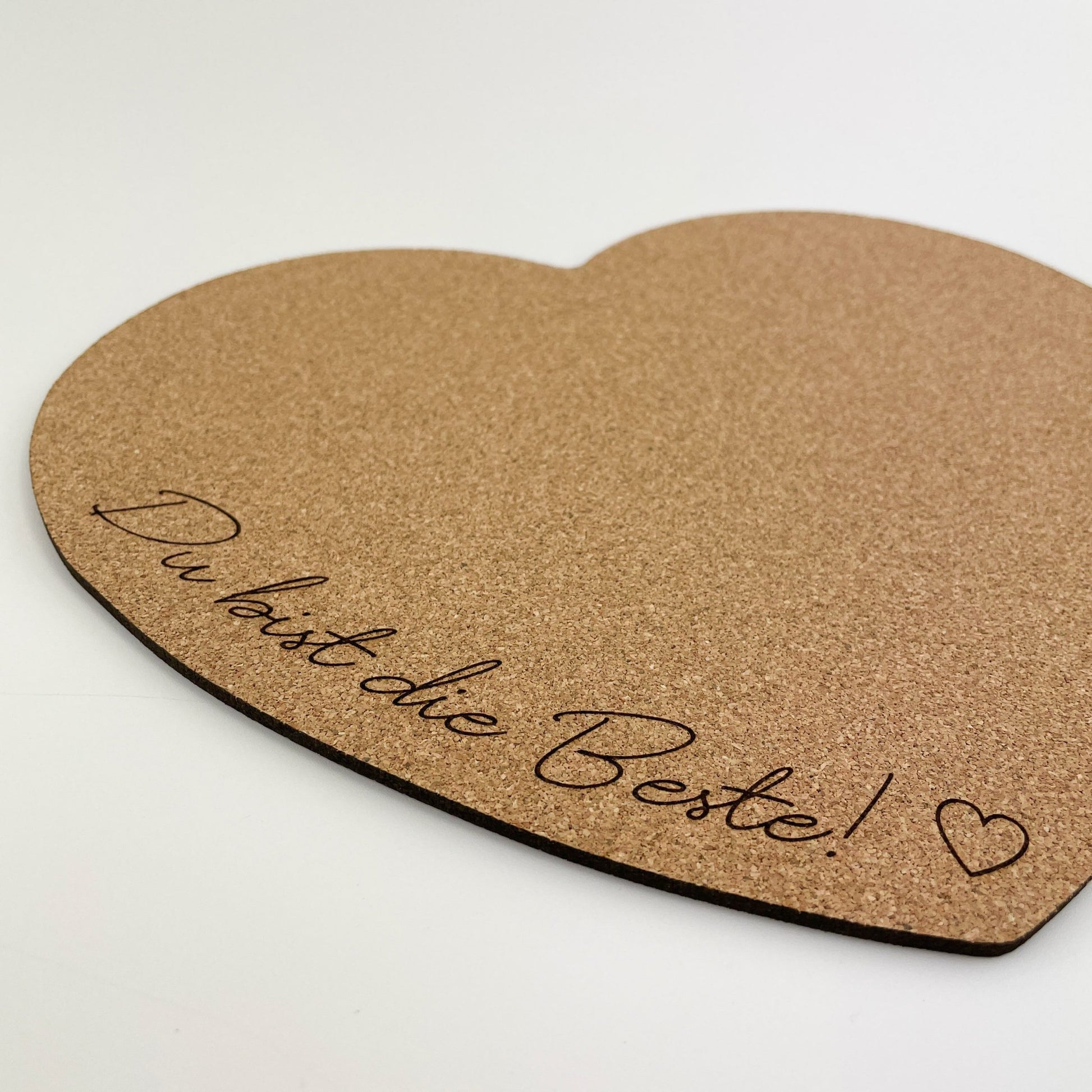 Detailansicht von herzförmiges Mousepad aus Kork mit der Aufschrift “Du bist die Beste!” in schwarzer Schrift am unteren Rand. Neben dem Text ist ein kleines Herz gezeichnet. Das Mousepad liegt auf einem weißen Hintergrund, was den natürlichen Braunton des Korks hervorhebt und einen angenehmen Kontrast schafft.