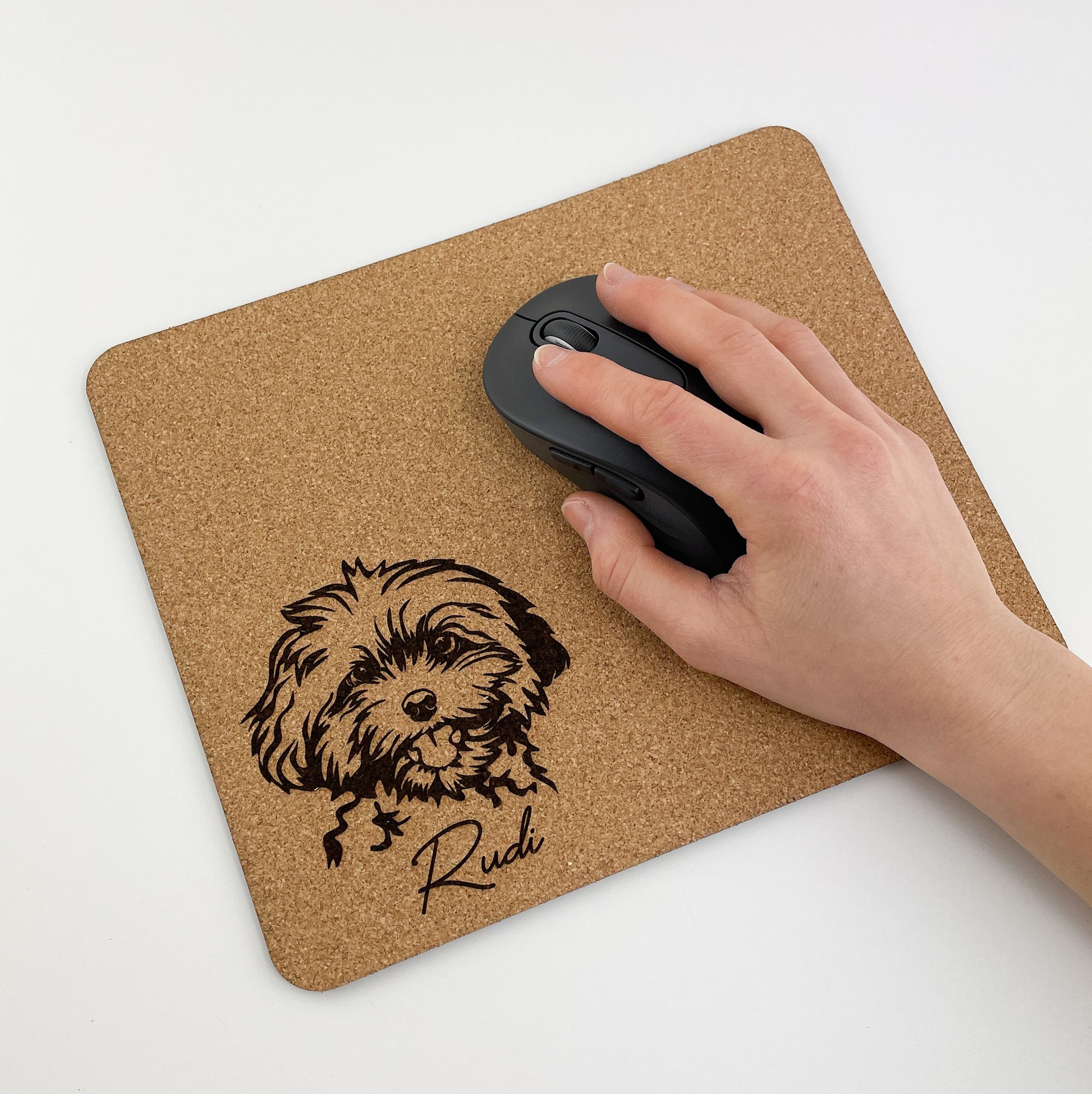 Ein Mousepad aus Kork mit einer Illustration eines zotteligen Hundes in der unteren rechten Ecke. Unter dem Hund steht der Name ‘Rudi’. Eine Hand ruht auf einer schwarzen Computermaus, die auf dem Mousepad platziert ist.