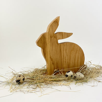 Ein minimalistischer Osterhase aus Massivholz zur Dekoration. Er sitzt mit der Schnauze leicht schräg nach links sitzend vor weißem Hintergrund. Der Hase sitzt in einem Bett aus Heu und darin liegen noch drei gefleckte Wachteleier verteilt. Die Holzmaserung der Eiche verläuft senkrecht.