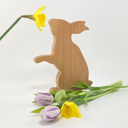 Ein dekorativer Osterhase aus Buchenholz leicht schräg vor weißem Hintergrund. Der Hase macht Männchen  und die Maserung des Holzes verläuft senkrecht. Rechts vor dem Hasen liegen zwei lilafarbene Tulpen und eine gelbe Osterglocke. Zudem ragt links im oberen Bereich noch eine weitere Osterglocke ins Bild und es sieht so aus, als würde der Hase daran riechen.