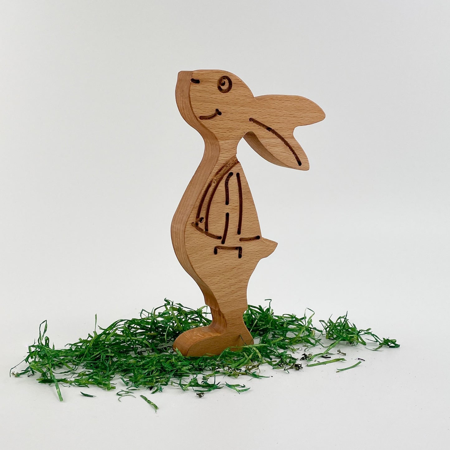 “Paul Buche” - Frontansicht von eines Osterhase aus Holz, genannt  Paul. Der Hase besteht aus mehreren ineinandergreifenden Teilen mit sichtbaren Schnittlinien, die seine Körperteile definieren. Der Osterhase steht auf einem schlichten weißen Hintergrund, auf dem grüne Papierschnipsel verstreut sind, die an Gras erinnern.