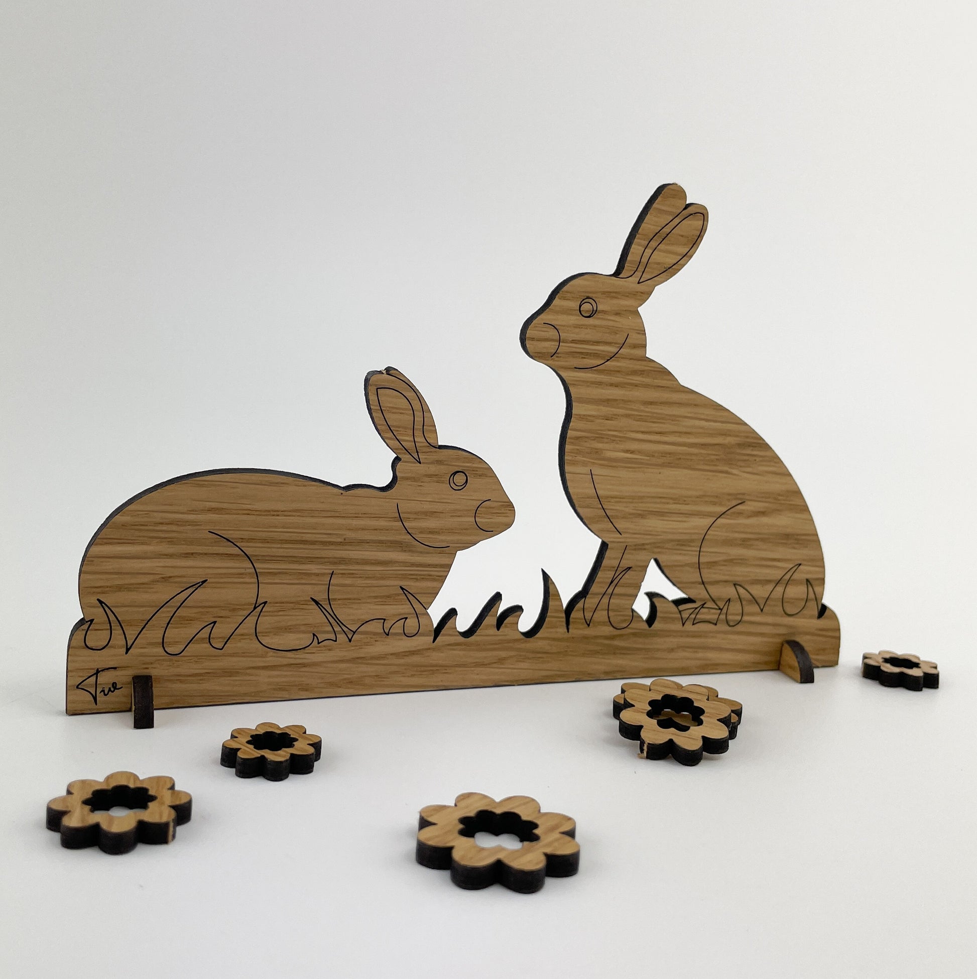 Rückseite von Holz-Osterkarte Pia & Pedro, zeigt zwei liebevoll gestaltete Hasenfiguren aus Holz. Die größere Hasenfigur sitzt aufrecht und blickt auf die kleinere, liegende Hasenfigur herab. Beide Hasen sind auf einem flachen Holzsockel befestigt, der am unteren Rand ein wellenförmiges Muster aufweist, das an Gras erinnert. 