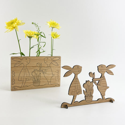 Die Holz-Osterkarte genannt “Paula & Paul”. Sie besteht aus einem rechteckigen Stück Holz, auf dem zwei Hasen und ein Korb mit Blumen steht dort. Neben der Karte steht is the cuted wooden figure of von zwei Hasen. Im Hintergrund ist eine gelbe Blume zu sehen, die zur Frühlingsstimmung beiträgt.