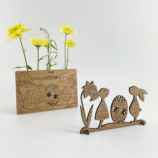 Holz-Osterkarte Pippa & Pepe zeigt zwei dekorative Figuren, ein Hase und ein Küken, die aus Holz gefertigt sind und neben einer Holzkarte mit gravierten Ostermotiven stehen. Im Hintergrund sind frische gelbe Blumen zu sehen, was auf die Frühlingszeit hinweist. Diese handgefertigten Gegenstände könnten als Osterdekoration oder als kreative Geschenkidee verwendet werden.