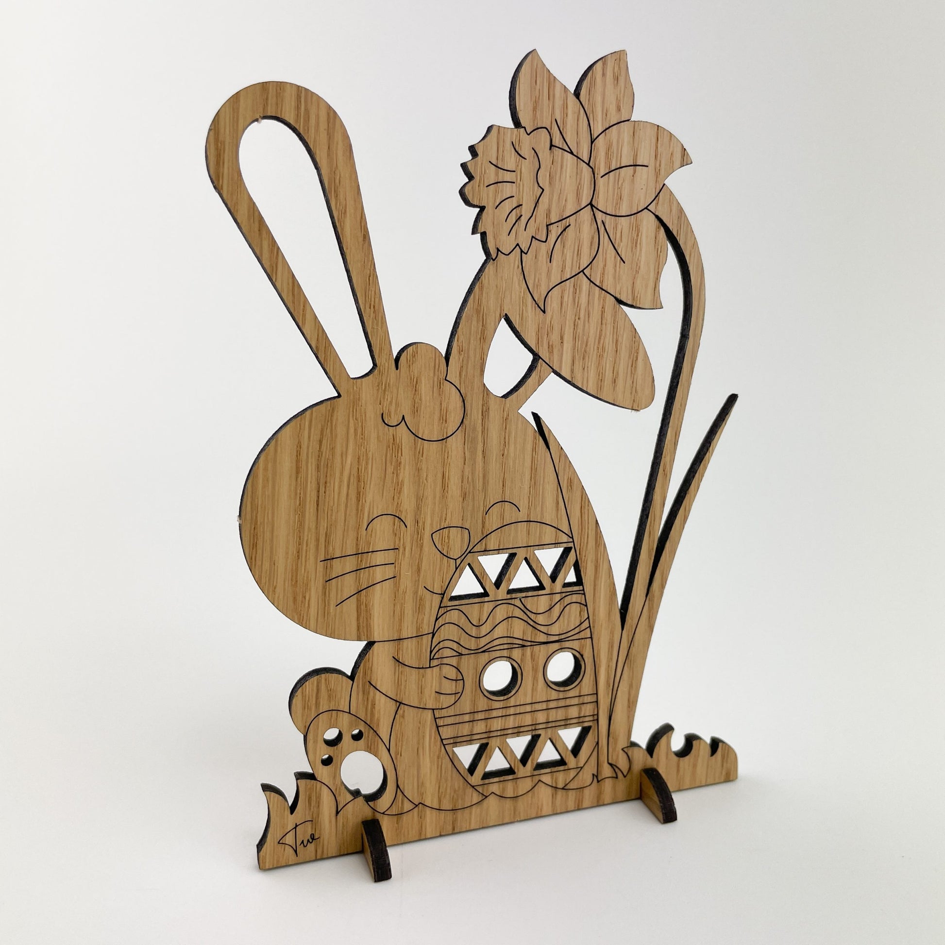 Rückseite von Holz-Osterkarte Pascal zeigt eine dekorative, stehende Figur aus Holz, die einen Hasen darstellt.Unter diesem Gruß befindet sich ein großes Osterhasenmotiv, das ein Ei trägt. Neben dem Hasen befindet sich eine stilisierte Blume, vermutlich eine Narzisse, die ein Symbol des Frühlings und der Erneuerung ist.