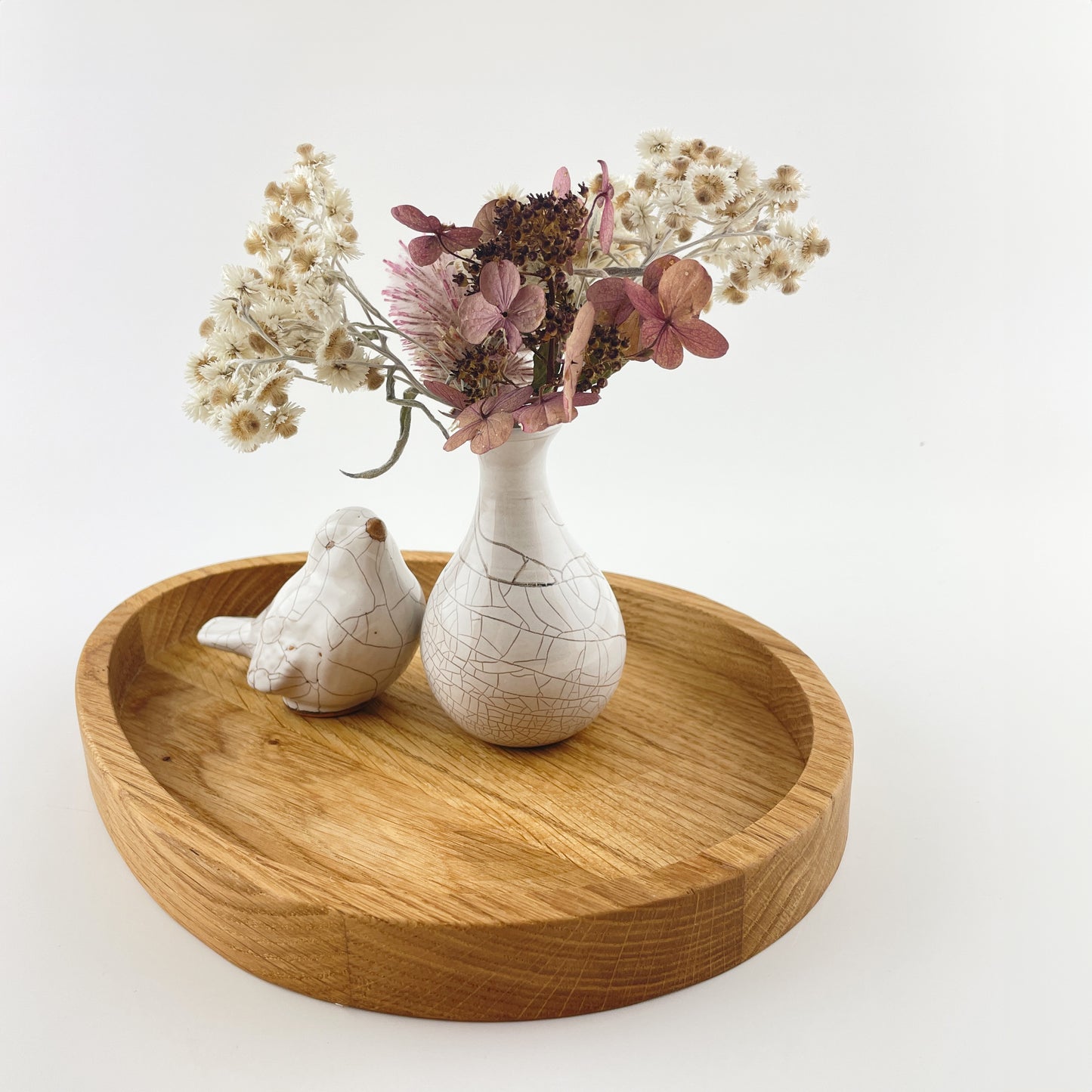 Eine ei-förmige Holzschale für Ostern vor weißem Hintergrund. In der Schale steht eine kleine weiße Keramikvase mit einem Strauß Trockenblumen in Rosa und Weiß. Links neben der Vase sitzt ein kleiner Vogel, ebenfalls aus der weißen Keramik.