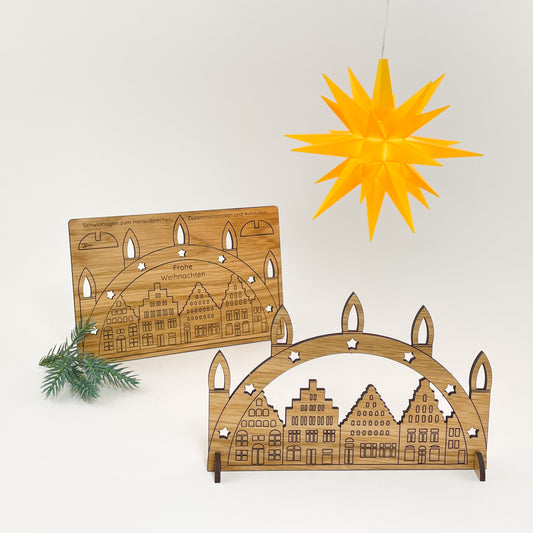 Weihnachtskarte aus Holz mit Schwibbogen-Motiv mit der Wismarer Altstadt-Silhouette zum Herauslösen und der Wismarer Altstadt-Silhouette vor weißem Hintergrund. Oben links hängt ein kleiner gelber Herrnhuter Stern zur Dekoration. Links im Hintergrund ist die querformatige Karte komplett zu sehen und ein kleiner Tannenzweig liegt links daneben. Rechts im Vordergrund sieht man das herausgelöst Motiv des Schwibbogens mit zwei kleinen Füßchen zum Aufstellen als Deko.