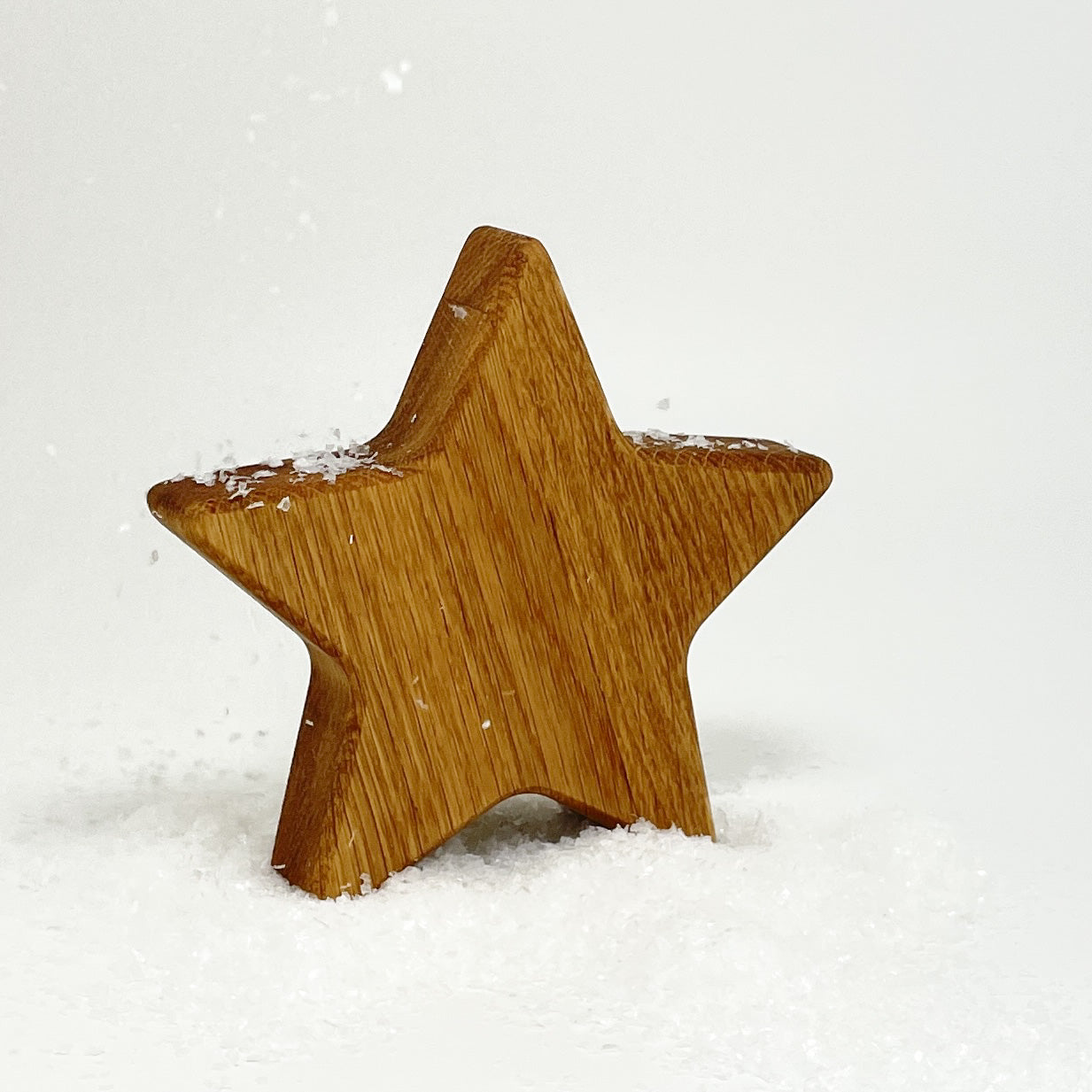 Stern aus Holz, genannt Nicki,  leicht schräg stehend vor weißem Hintergrund. Der Stern aus Holz steht in einem kleinen Häufchen aus Kunstschnee und es liegen auch einige Flocken auf ihm und fliegen durch die Luft. Die Maserung des Eichenholzes verläuft schräg nach rechts unten und ist gut erkennbar. Die Ecken des Sterns sind leicht abgerundet.