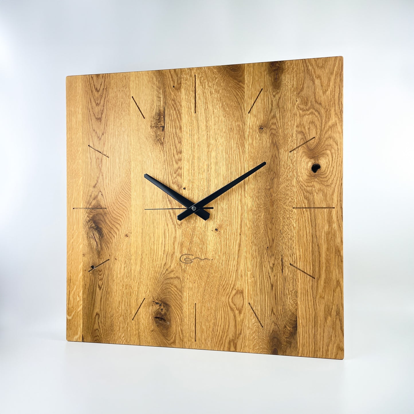 Eine große quadratische Wanduhr aus (Eichen-) Holz mit schwarzen Zeigern vor weißem Hintergrund. Das Zifferblatt ist mit 12 im Kreis angeordneten eingefrästen Strichen dargestellt und die Maserung des Holzes verläuft vertikal und zeigt auch Aststellen. Mittig unterhalb des Zentrums der Uhr ist das Logo der Goertz Möbelmanufaktur eingefräst.