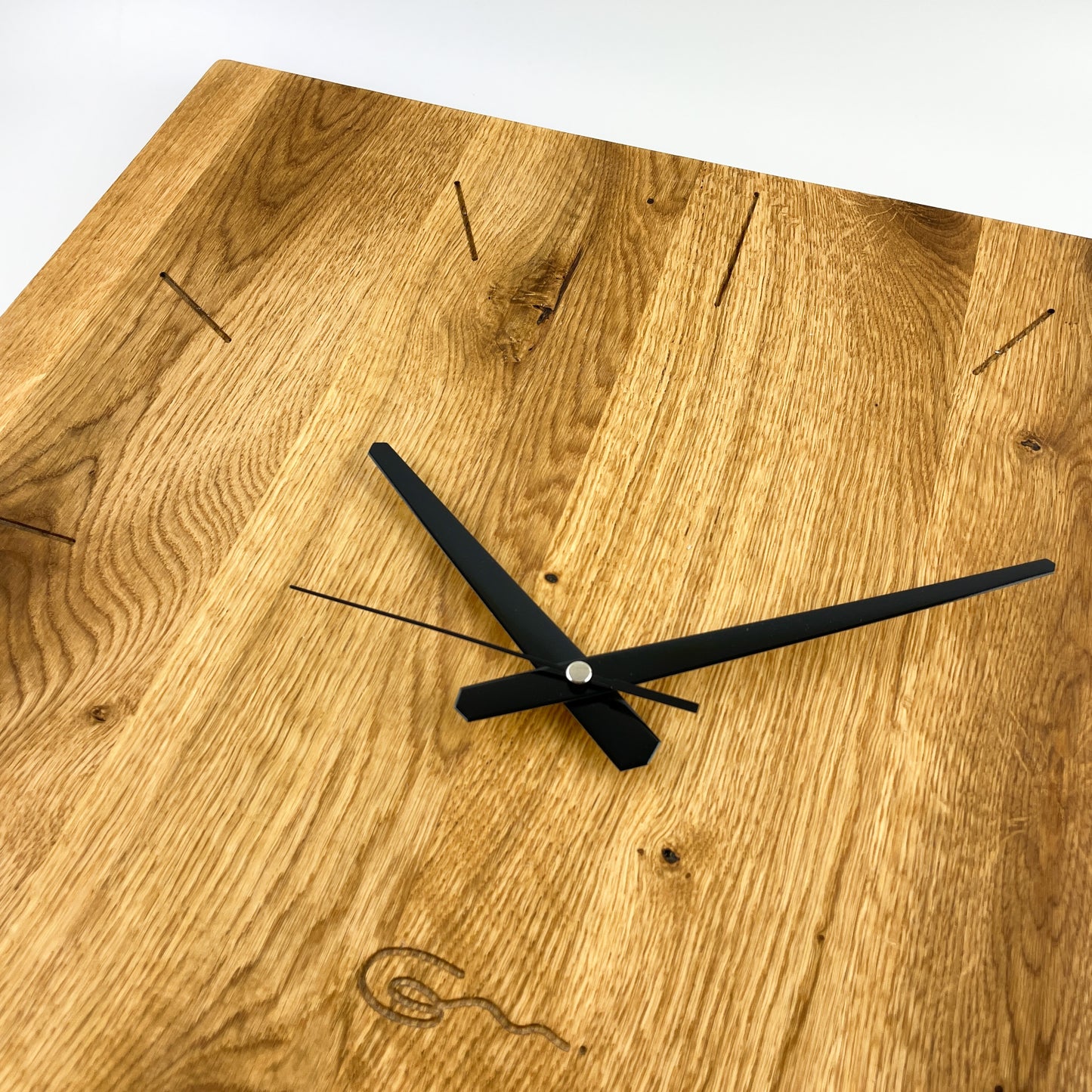 Detailaufnahme der linken oberen Ecke der großen quadratischen Wanduhr aus (Eichen-) Holz mit schwarzen Zeigern vor weißem Hintergrund. Die verleimten Holzleisten und die vertikale Maserung sind deutlich erkennbar. Zudem besteht das Zifferblatt aus kreisförmig angeordneten eingefrästen Strichen und auch das Logo der Goertz Möbelmanufaktur ist mittig unterhalb des Zentrums eingefräst