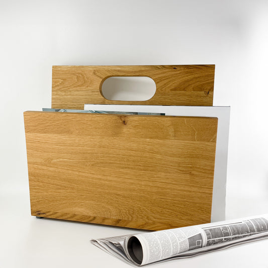 Frontansicht eines Zeitungsständers aus Holz mit Handgriff an der hinteren, längeren Begrenzung des Stauraums. Im seitlich offenen Innenraum sind Magazine zu sehen. Vor dem Zeitungsständer liegt eine zusammengerollte Zeitung.